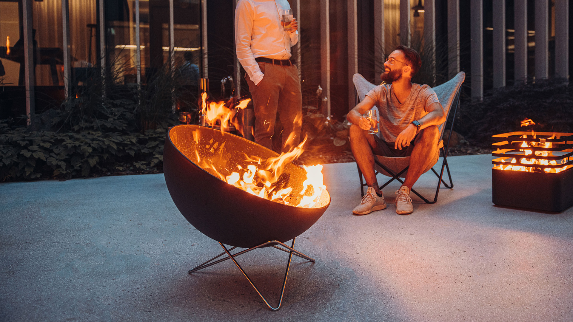 The 00259 Bowl 70 Star Stand integrates seamlessly into the setting, where a man and woman sitting in chairs around a fire pit.