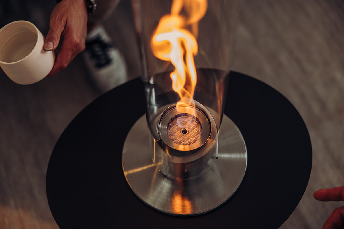 A person is holding the 00696 Spin 1200 Bio Burner placing it in the SPIN Tablefire, adding a functional and elegant touch to the scene.