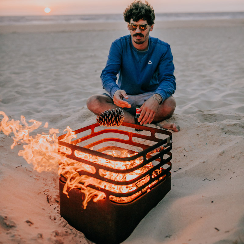 A man sitting on the beach with a fire, with the Cube Firebasket Rusty adding a functional and elegant touch to the scene.