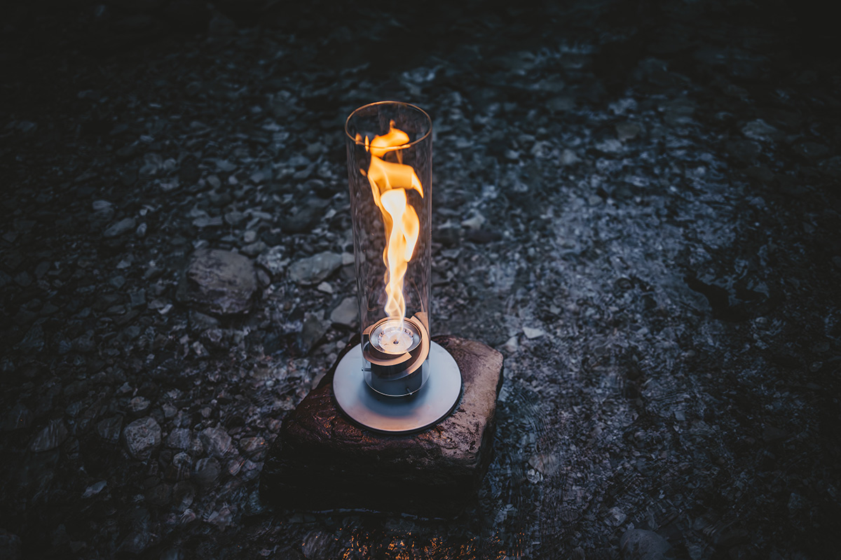 SPIN 1200 Tabletop Fireplace grey A Tablefire is lit on a rock, with the 00697 Spin 1200 Tablefire grey adding a functional and elegant touch to the scene.