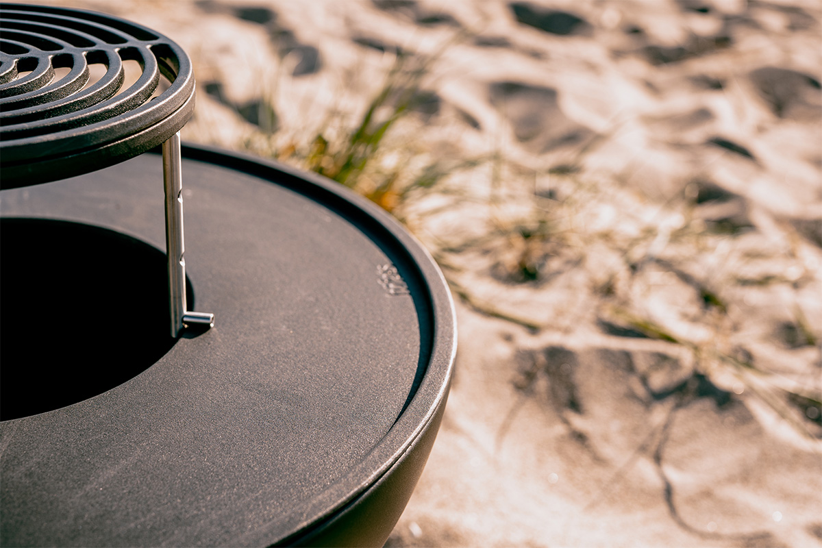 Illustrated is a black fire pit sitting on a sandy beach, with the BOWL 57 Warming Ring.