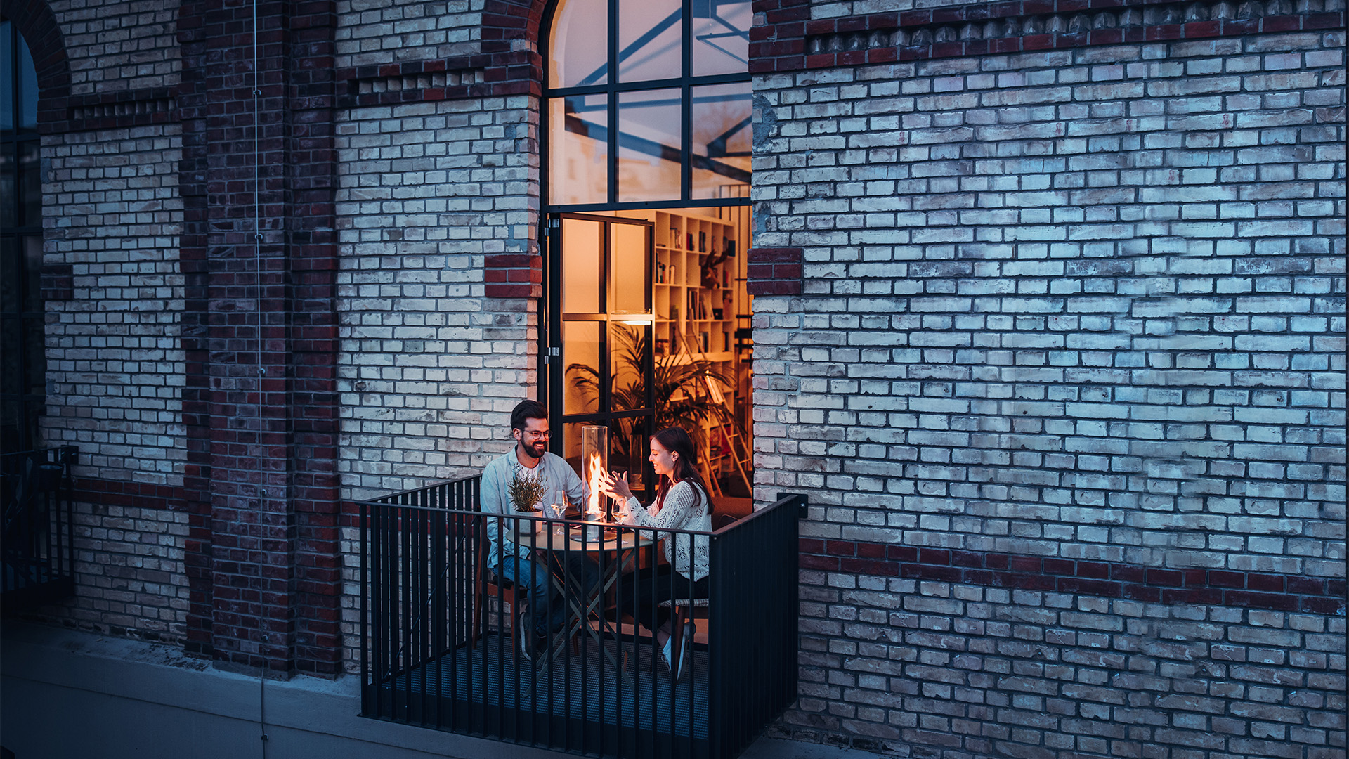 Illustrated is a man and woman sitting on a balcony looking out a window, with the SPIN 1200 Tabletop Fireplace silver.