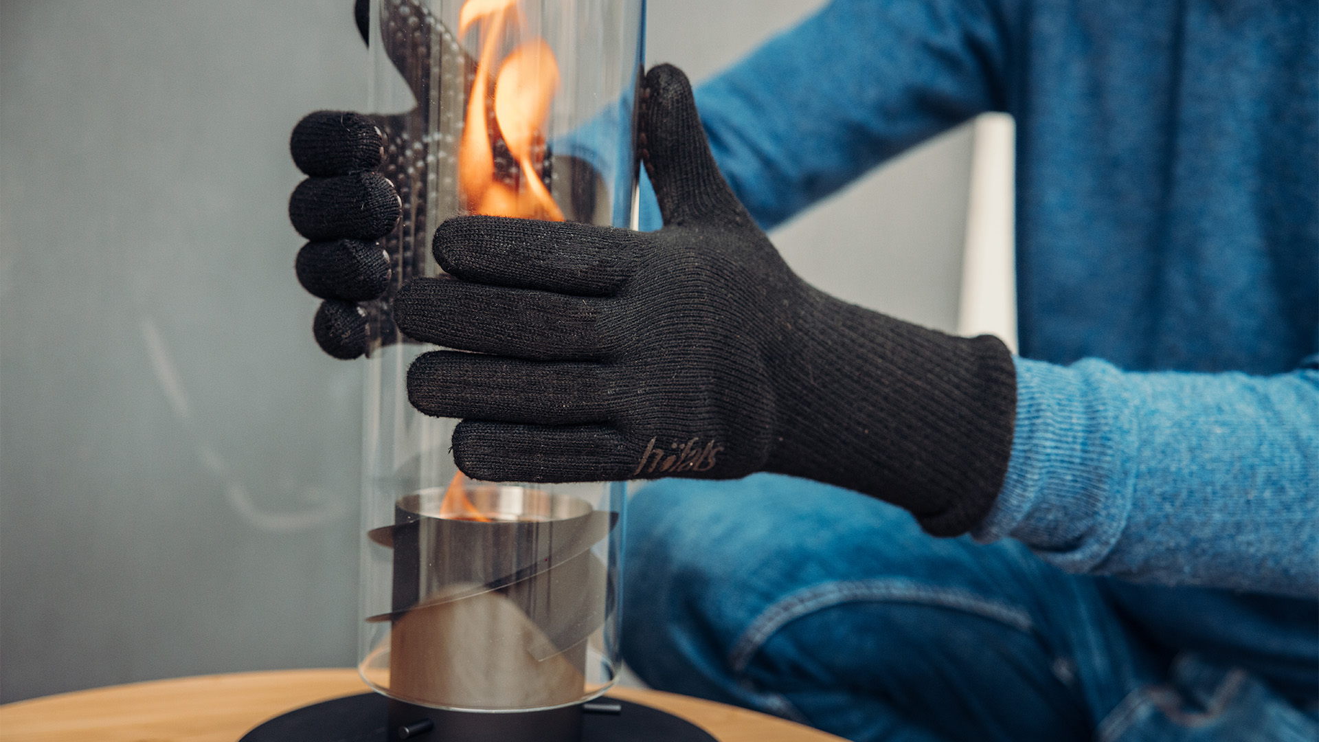 This image shows a person in a blue sweater and black gloves holding a tabletop fireplace with a fire inside, with the Fire Gloves Aramid.