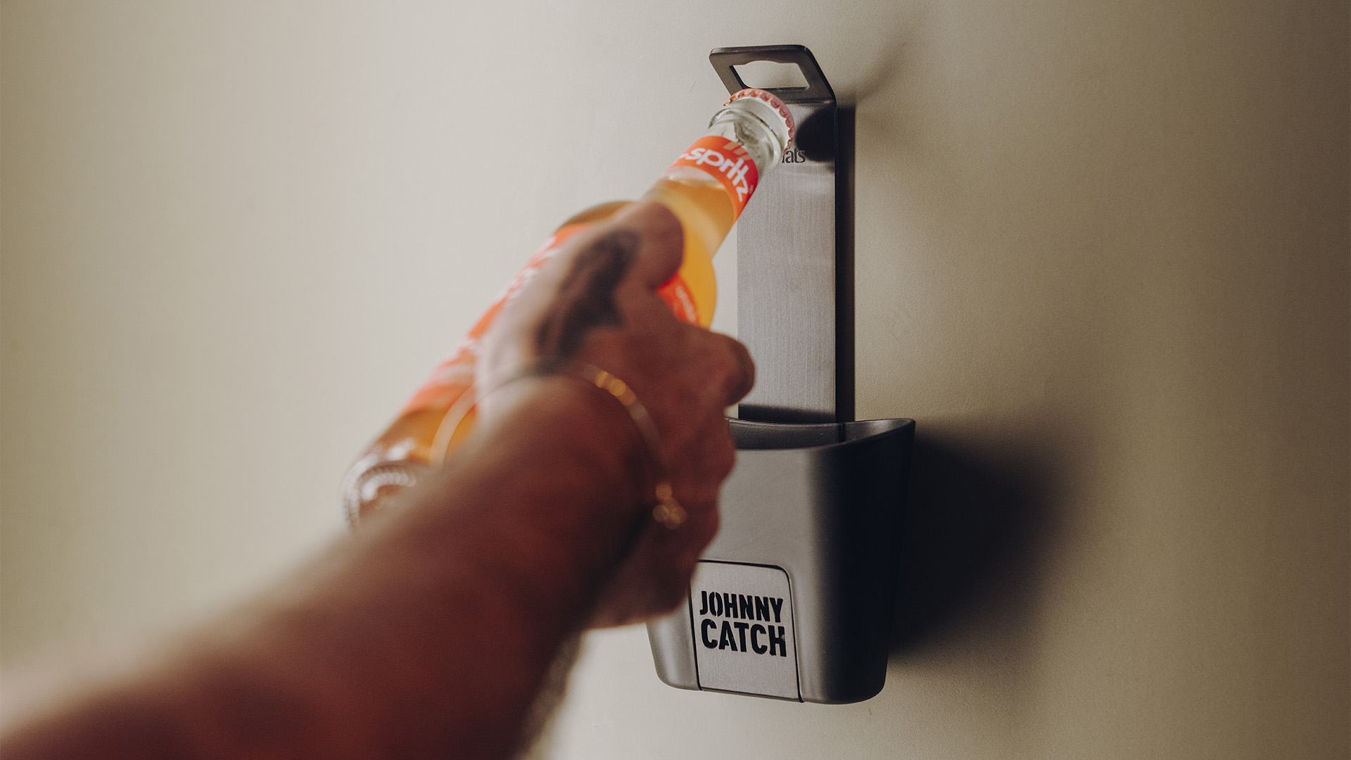 A depiction of a hand holding a bottle of orange juice which is about to be opened with a bottle opener, featuring the 00842 Johnny Catch Cup Black Bottleopener as a central and purposeful element.