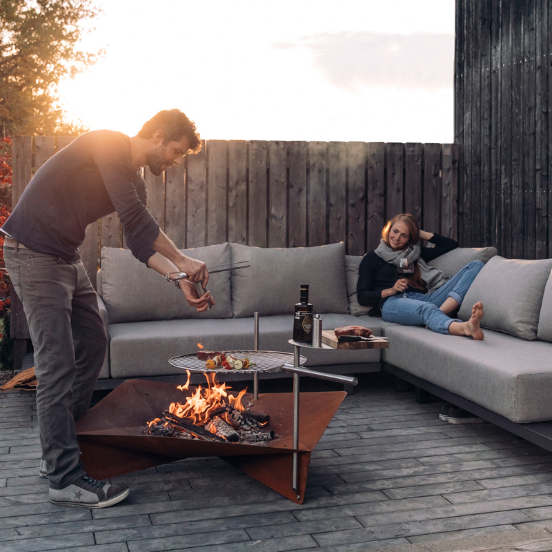 A man is setting a fire in a fire pit, enriched by the presence of the Triple Fireplace, which merges design with utility.