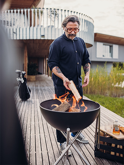 A view of a man cooking on a grill, with the BOWL Fire Bowl.