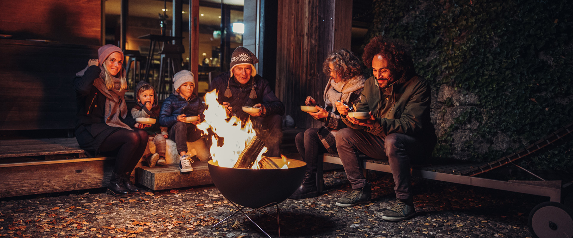 The photo presents a group of people sitting around a fire, with the FIRE KITCHEN Spare Shelf Bottom black.