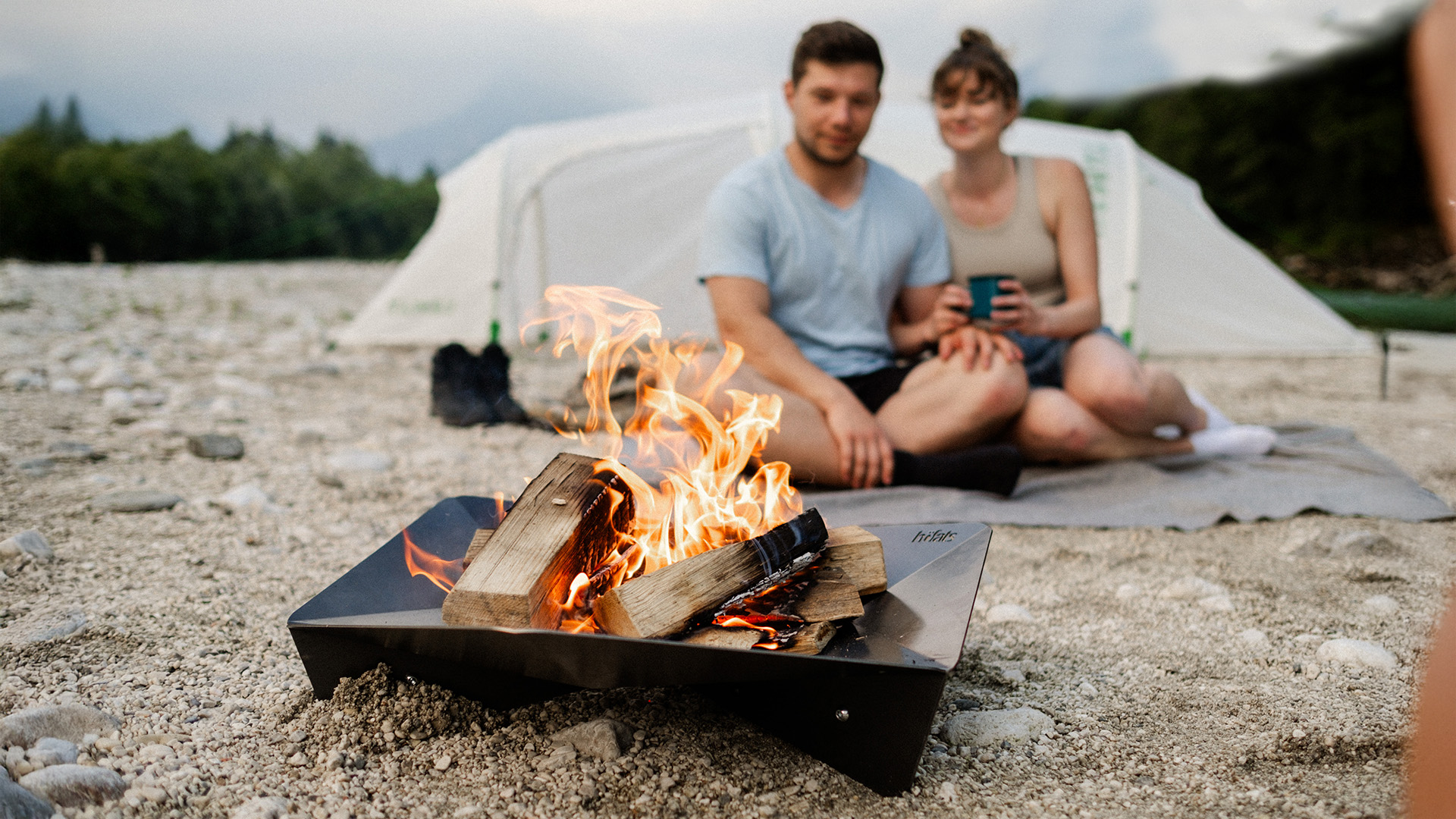 A man and woman sitting on a blanket by a campfire, with the Triple 65 Fireplace adding a functional and elegant touch to the scene.