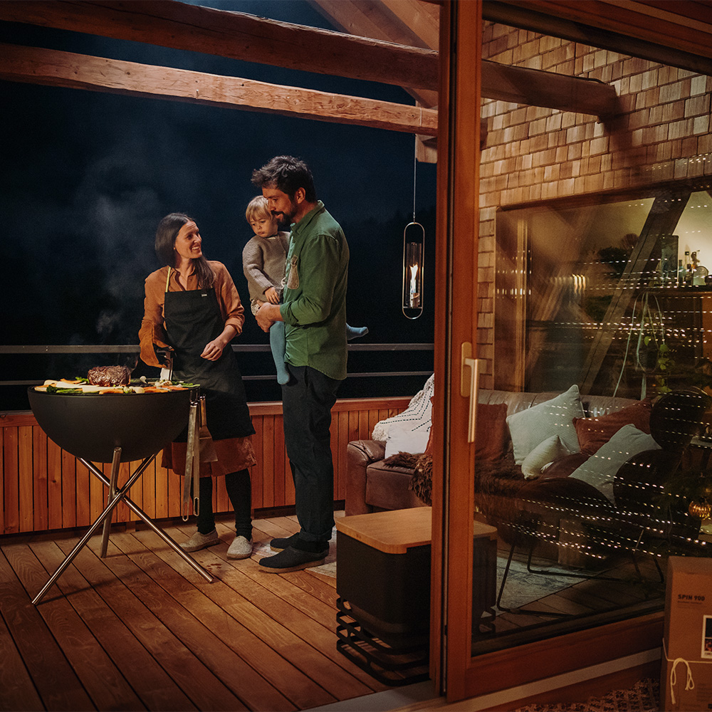 A man and woman are standing on a balcony using the BOWL 70 Firebowl cooking food.