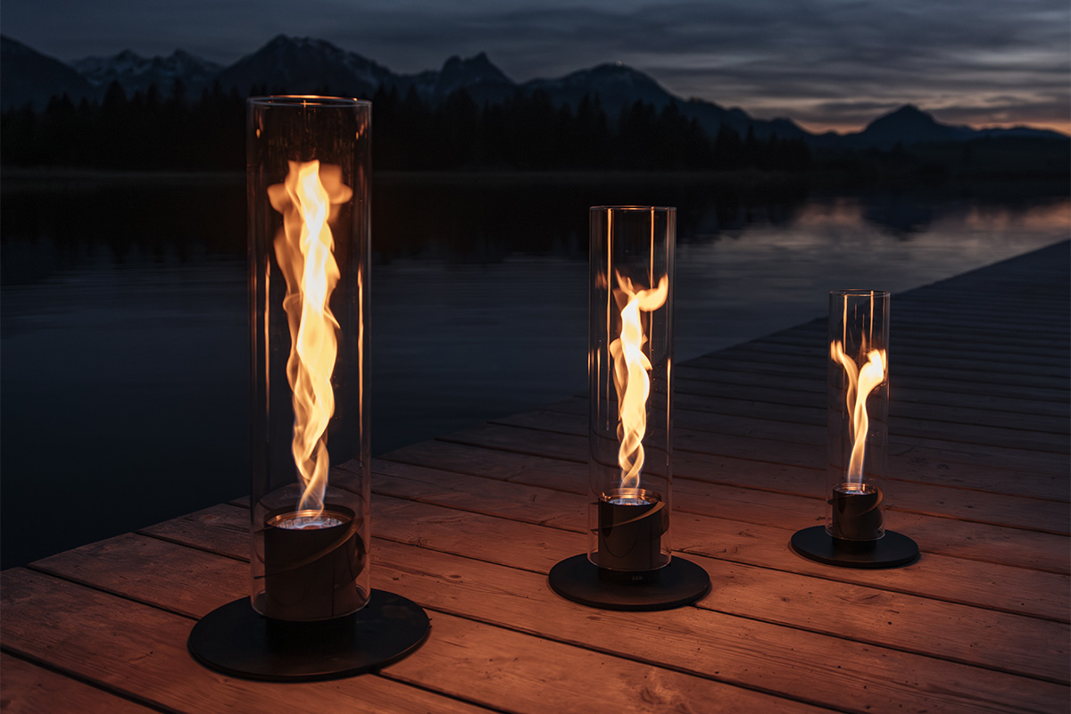 SPIN 1500 Pedestal Fireplace black The 00628 Spin 1500 Tablefire black integrates seamlessly into the setting, where two torches on a dock with a lake in the background.