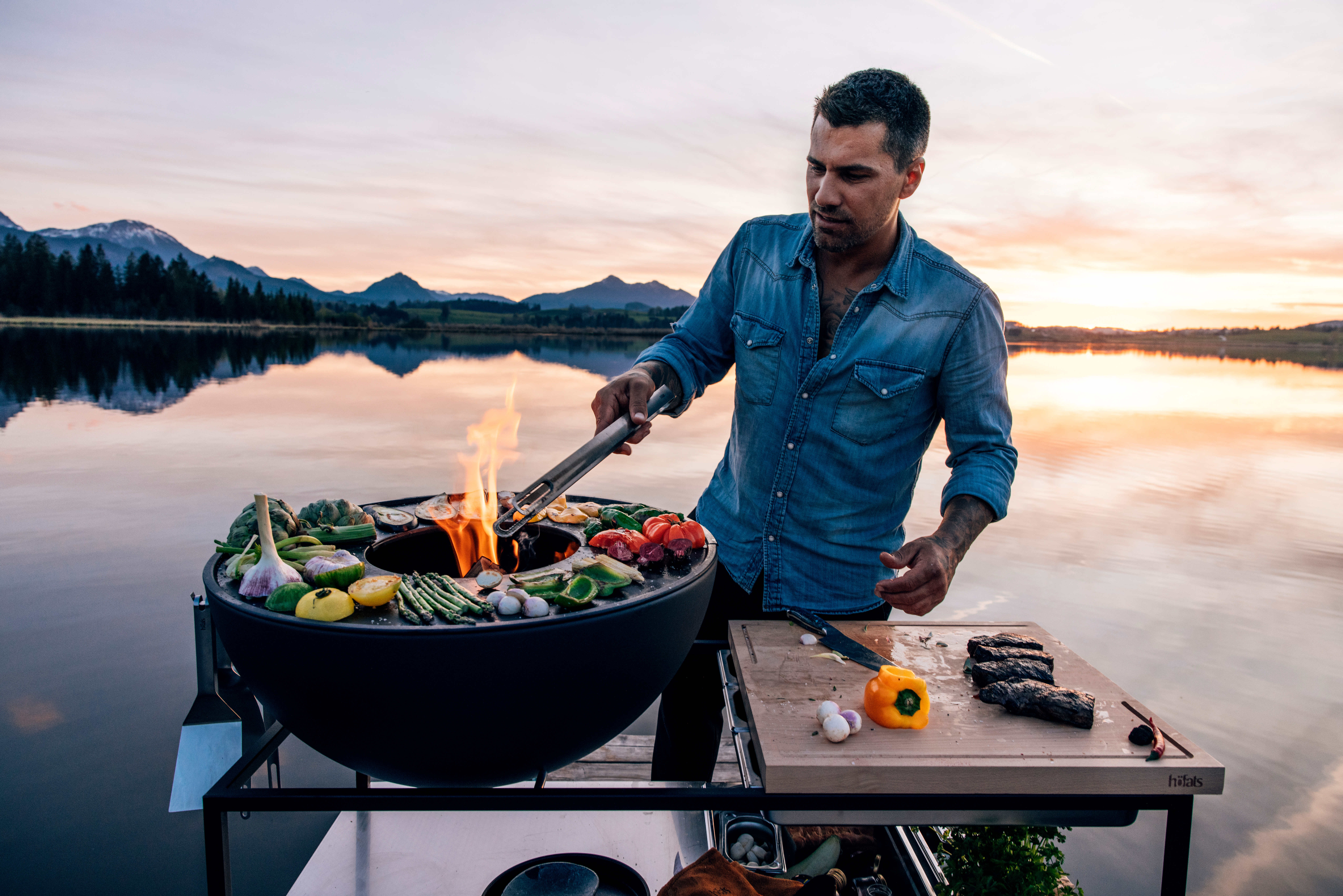 Illustrated is a man cooking on a grill on a lake, with the FIRE KITCHEN with BOWL 70 Plancha BBQ Set incl. wheels.
