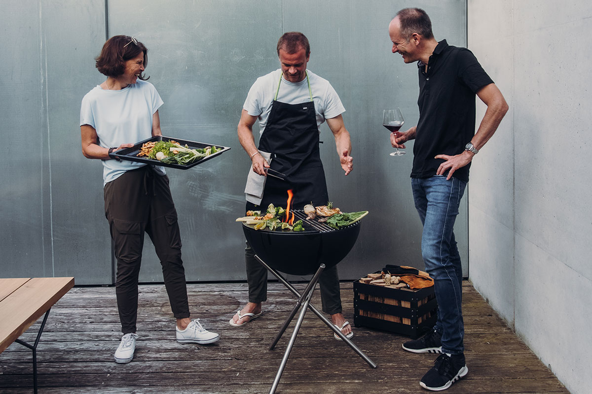 The photo presents a man and woman are cooking on a grill, with the BOWL 57 Fire Set flex.