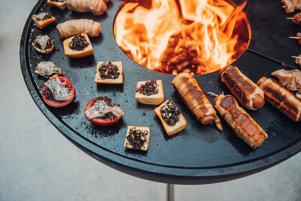 Illustrated is a fire pit with a bunch of meat on it, with the BOWL 70 Plancha BBQ Set high.