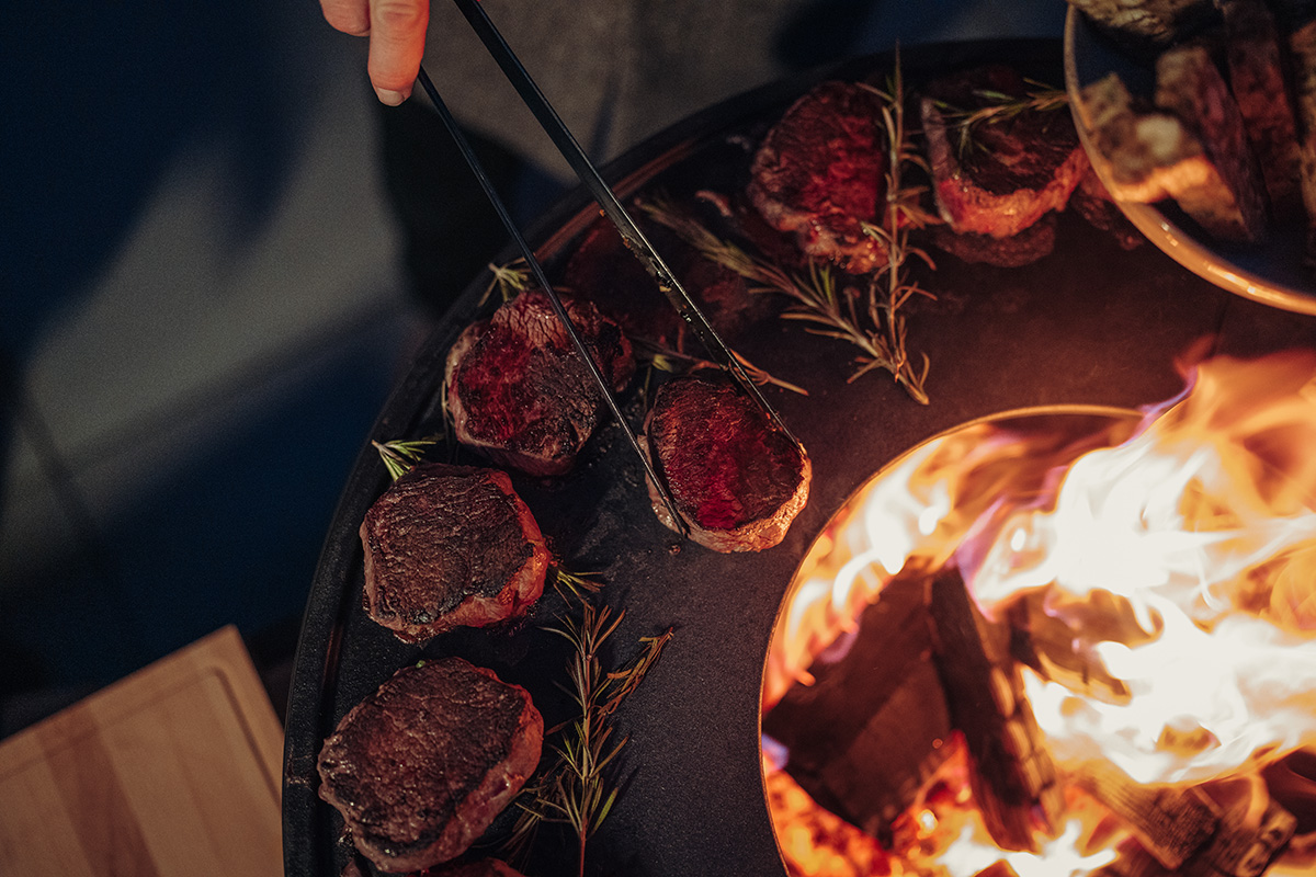 A person is cooking steaks on a grill, enriched by the presence of the 00859 Tong black, which merges design with utility.