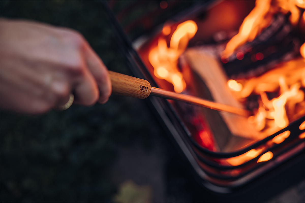 A person holding a wooden stick over a fire, enriched by the presence of the 00581 Blow poker, which merges design with utility.