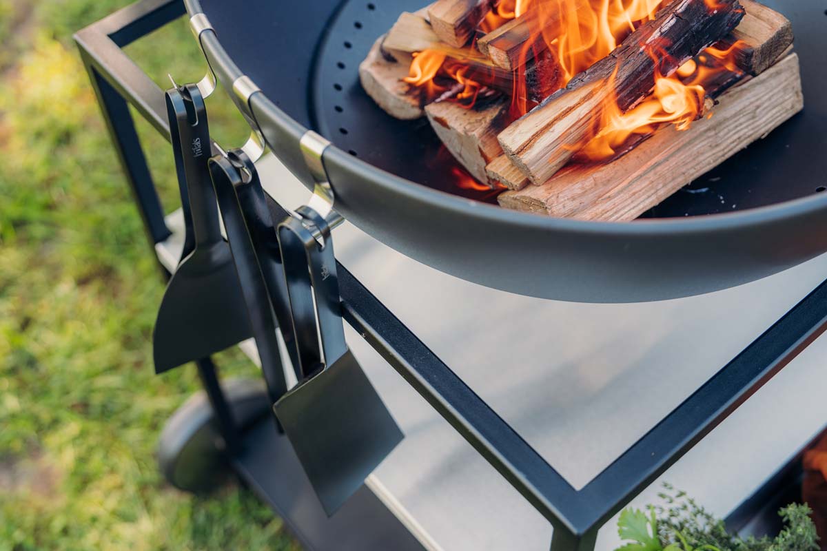 A depiction of a large metal fire pit with a firewood burning in it, featuring the 00878 Bbq Tool Set black as a central and purposeful element.
