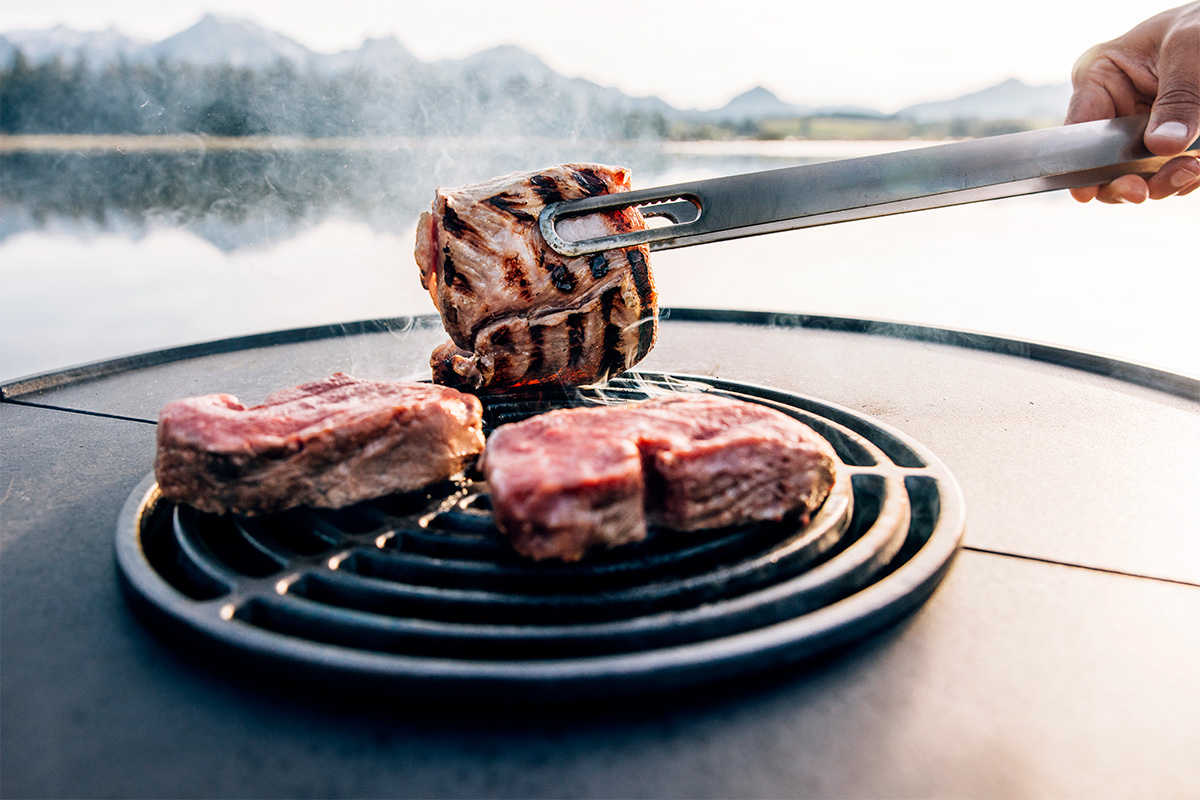 A person is grilling meat on a grill, enriched by the presence of the 00615 Bowl 70 Sear Grate, which merges design with utility.