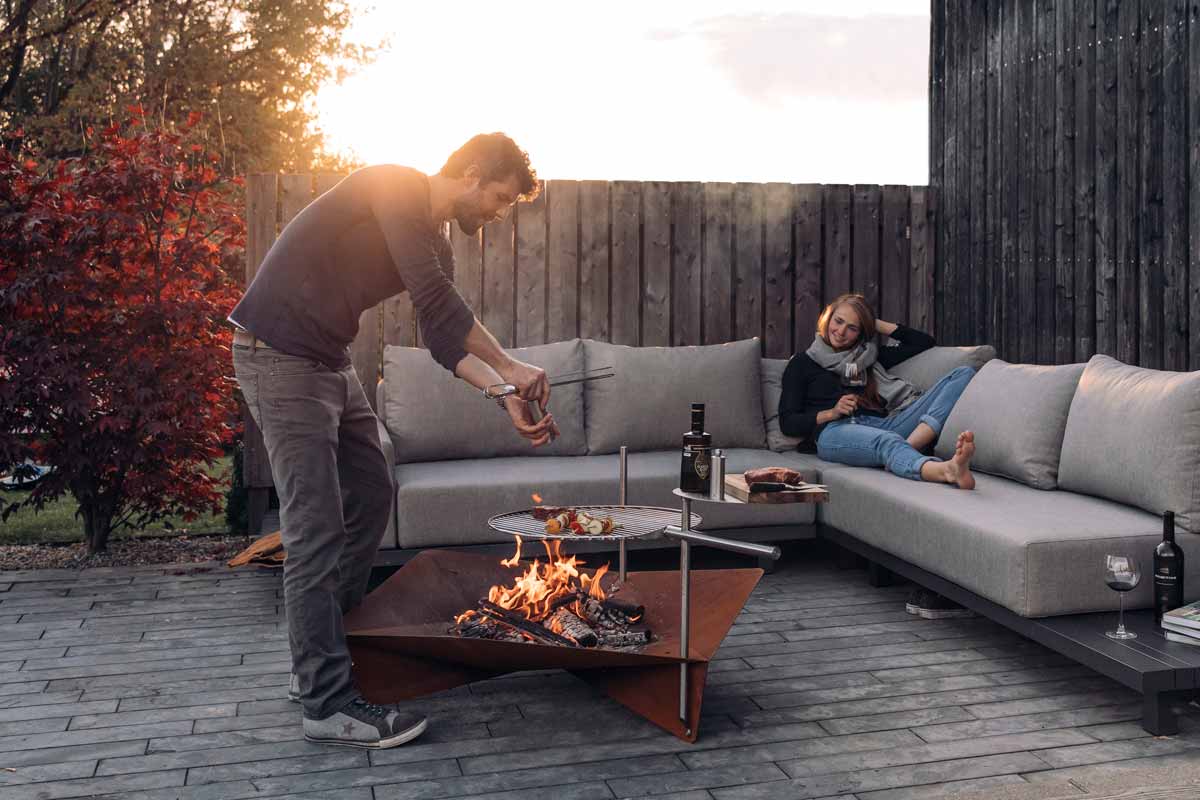 The photo presents a man and woman are sitting on a couch by a fire pit, with the TRIPLE 120 Fire Bowl.