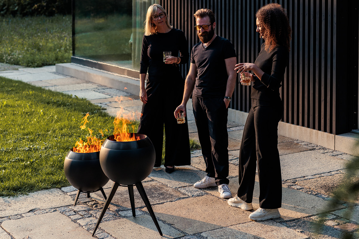 MOON 45 all black Fire Basket with high stand Drei schwarz gekleidete Personen stehen um zwei MOON Feuerschalen und unterhalten sich.