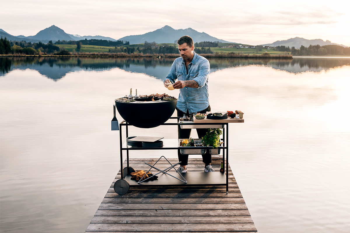 FIRE KITCHEN Stainless Steel Container Set (5 pcs) A man sitting on a dock with a grill on it, enriched by the presence of the 00764 FIRE KITCHEN Stainless Steel Container Set (5 pcs), which merges design with utility.