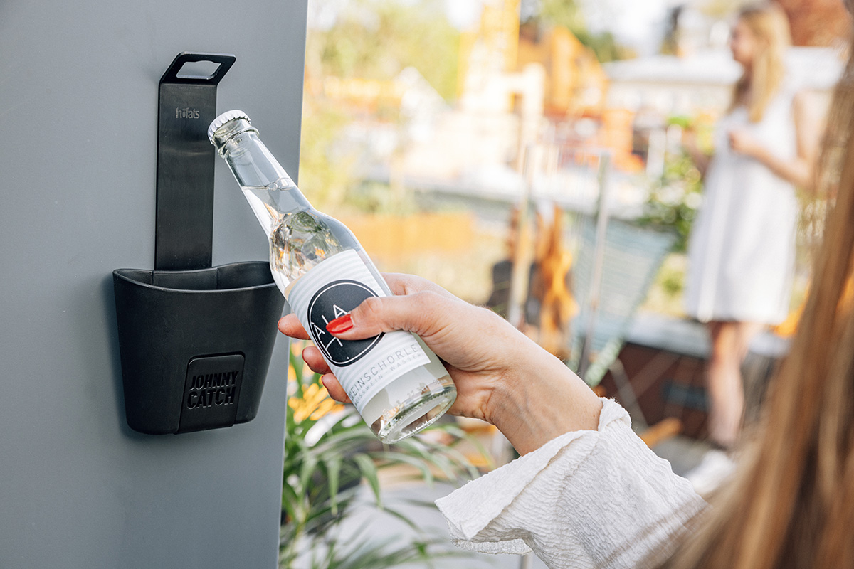 A depiction of a woman is opening a bottle of water, featuring the 00842 Johnny Catch Cup Black Bottleopener as a central and purposeful element.