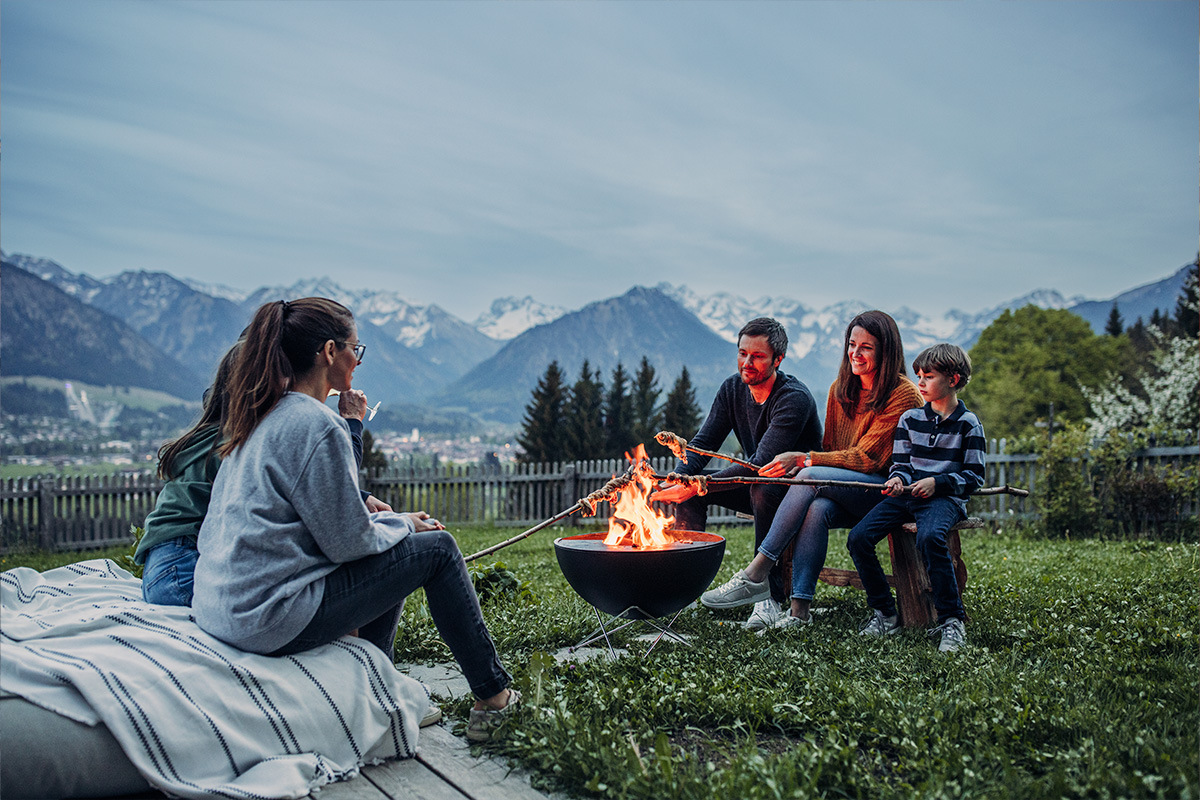 A woman and two children sitting around a fire pit, with the 00249 Bowl 57 with starstand adding a functional and elegant touch to the scene.