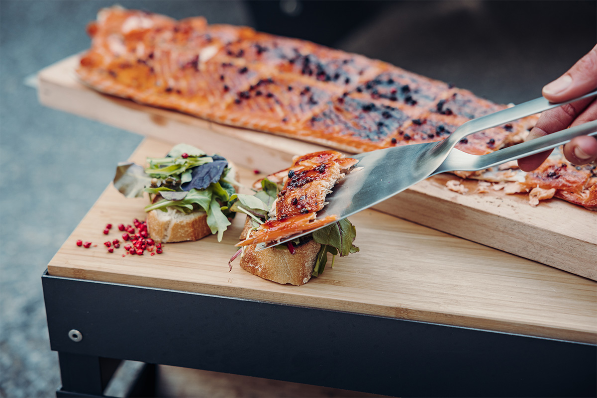 Flame-grilled salmon board The photo presents a person cutting a piece of pizza on a wooden cutting board, with the Flame-grilled salmon board.