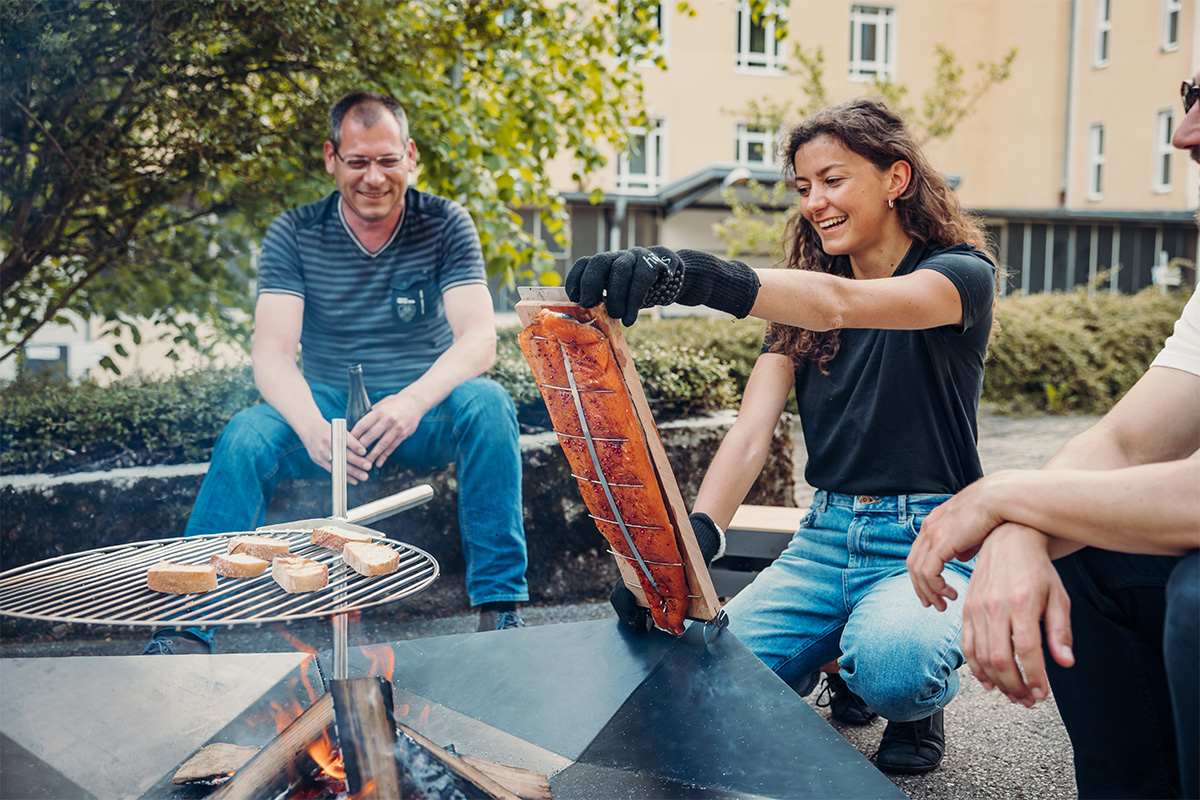 Flame-grilled salmon board Illustrated is a man and woman sitting around a fire pit, with the Flame-grilled salmon board.
