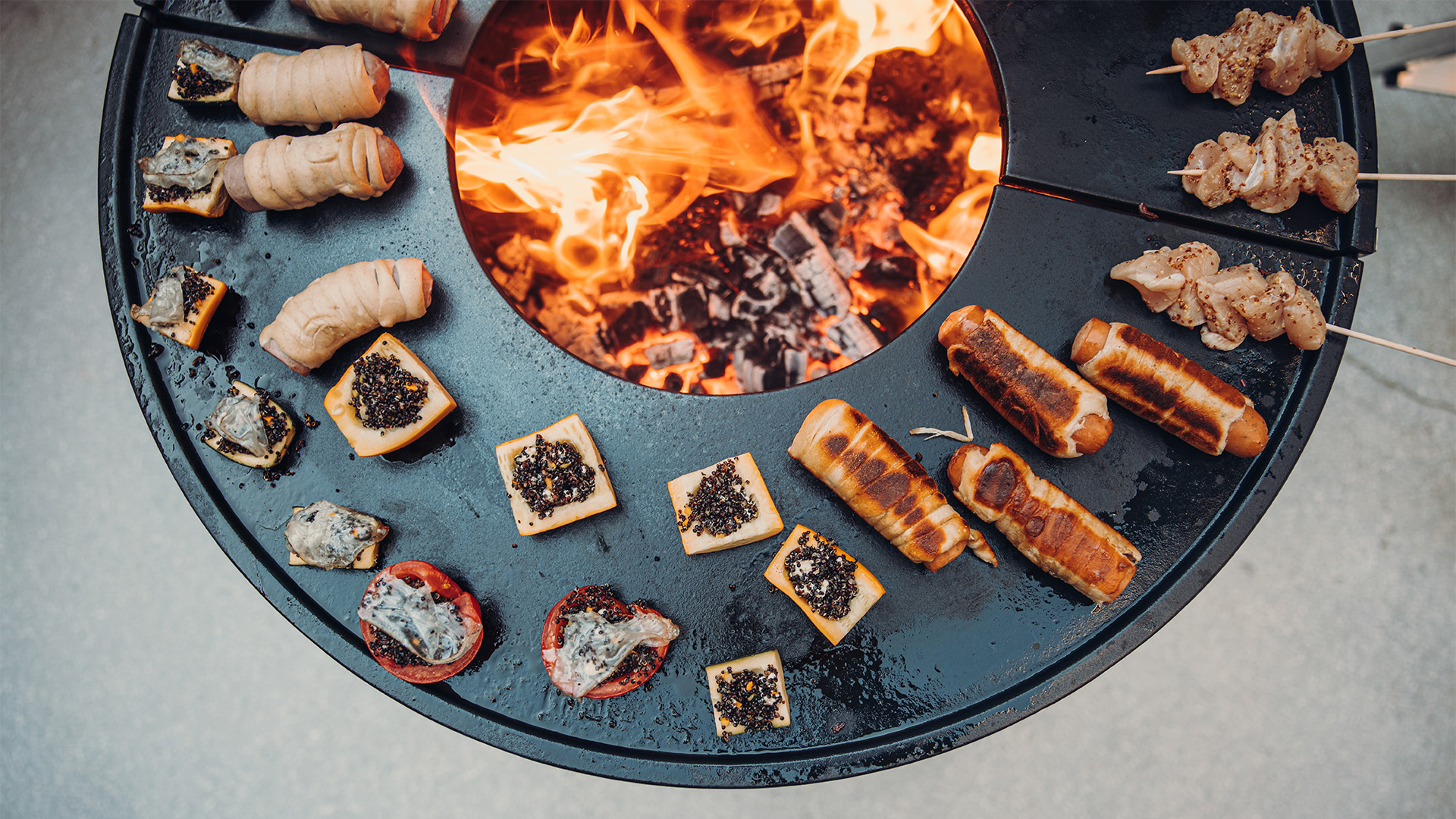 The photo presents a fire pit with a bunch of food on it, with the BOWL 70 Plancha BBQ Set low.
