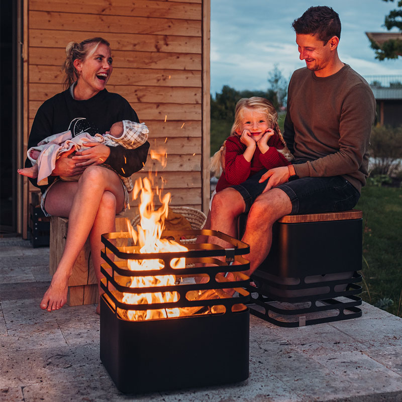 Experience a man and a woman sitting on a bench next to a fire with the refined touch of the Cube Firebasket