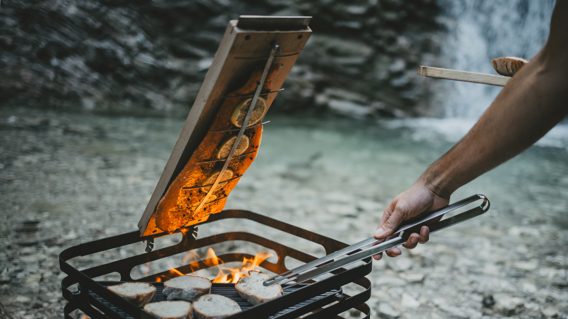 A depiction of a person holding a knife over a campfire, featuring the 00702 Flame-grilled salmon board3 as a central and purposeful element.