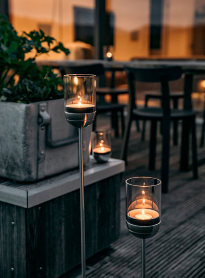 Depicted here is two candles on a wooden table with a plant in the background, with the LOGO GRAVITY CANDLE M90 Lantern.