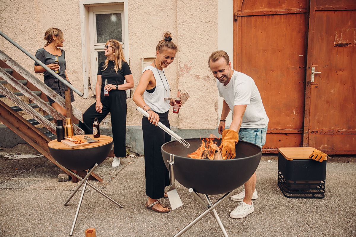 BOWL 70 Fire Bowl with tripod A group of people standing around a fire pit, with the 00258 Bowl 70 with Tripod adding a functional and elegant touch to the scene.