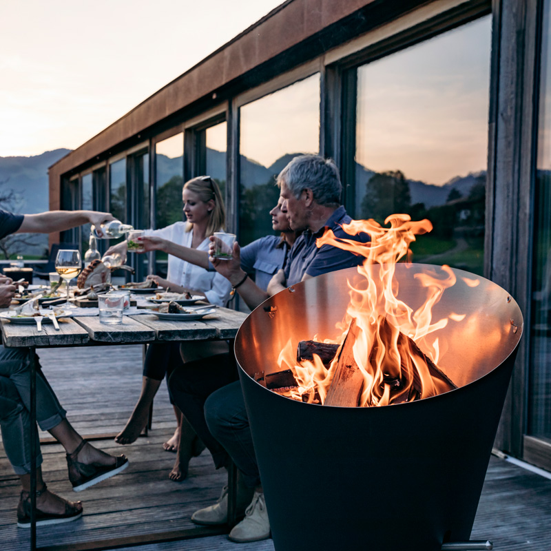 The photo presents a man and woman sitting at a table with a fire pit, with the CONE Charcoal Grill.
