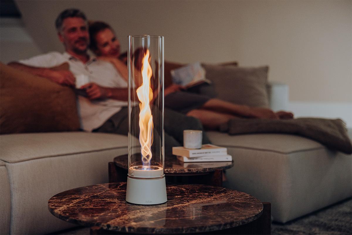 SPIN air 1200 creme steht auf einem Couch-Tisch im Hintergrund ist ein Sofa auf welchem zwei Personen liegen und in die Flamme schauen