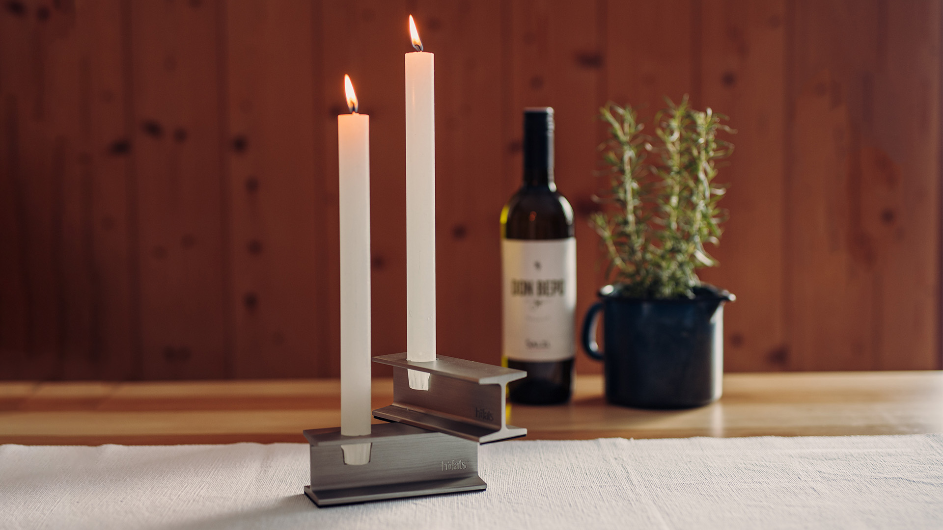The photo presents a candle and a bottle of wine on a table, with the HENRY METAL I Candleholder silver.