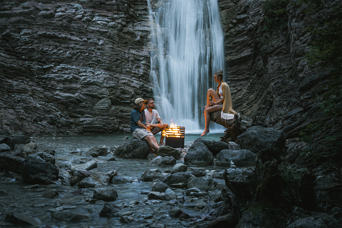 CUBE Fire Basket black The Cube Firebasket black integrates seamlessly into the setting, where two people sitting on rocks in front of a waterfall.