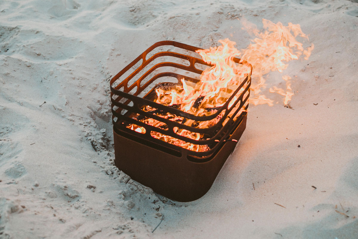 CUBE Fire Basket Corten Steel A fire pit is on the beach with sand, with the Cube Firebasket rusty adding a functional and elegant touch to the scene.