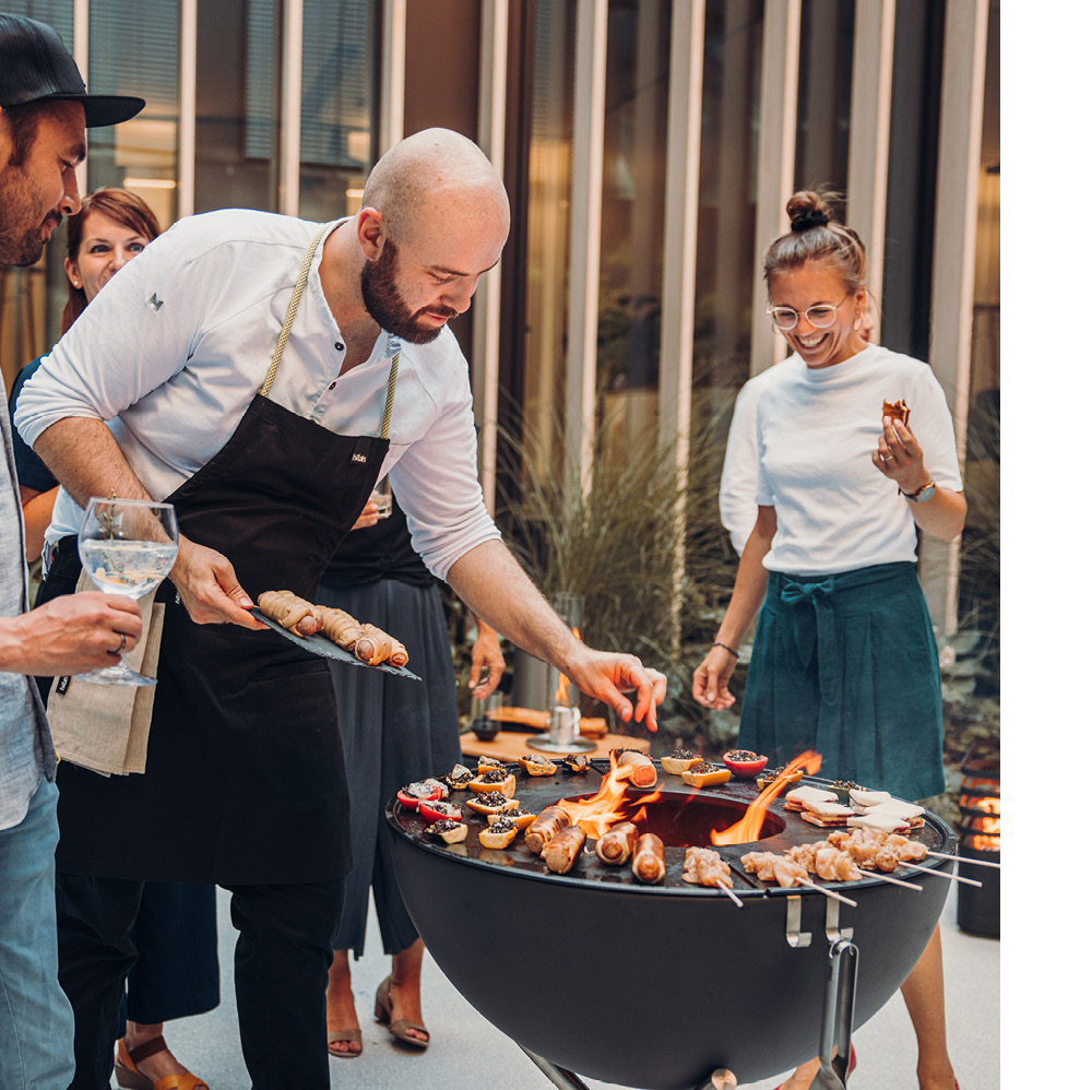 The photo presents a man cooking food on a grill while two women watch, with the CUBE Plancha.