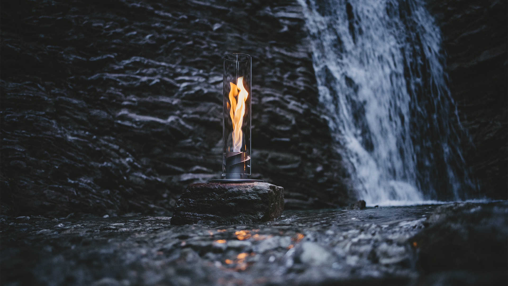A small fire is lit in front of a waterfall, with the 00697 Spin 1200 Tablefire grey adding a functional and elegant touch to the scene.