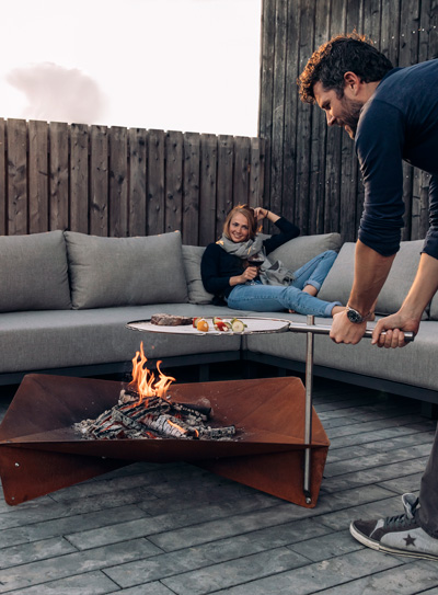 A depiction of a man and a little girl sitting on a couch by a fire pit, featuring the Triple Fireplace as a central and purposeful element.