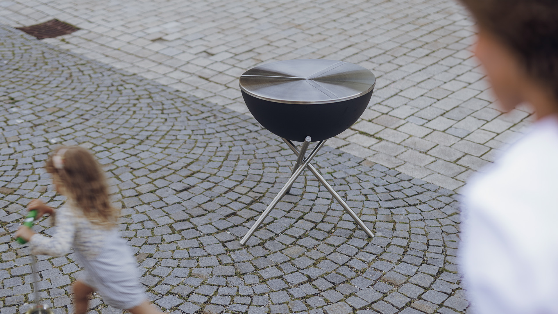 A depiction of a little girl running towards a grill, featuring the 00479 Bowl 70 Lid as a central and purposeful element.