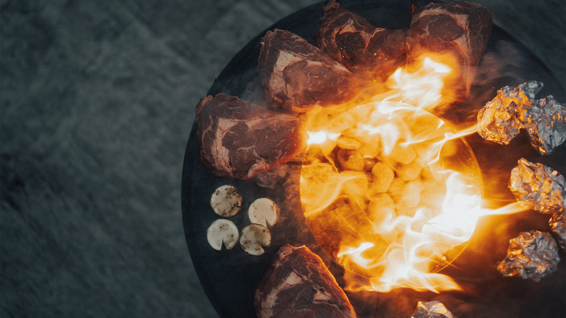 Depicted here is a fire pit with a bunch of meat on it, with the BOWL 57 Plancha BBQ Set low.