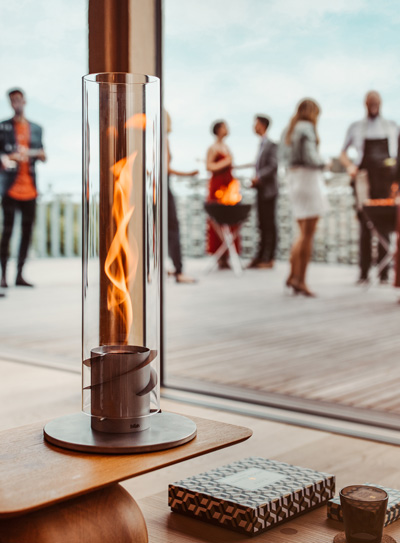 Depicted here is a fire is on the table in front of a glass door, with the SPIN 1200 Tabletop Fireplace silver.