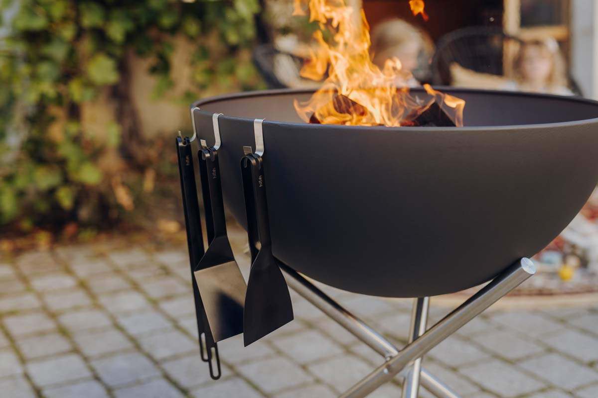 The 00878 Bbq Tool Set black integrates seamlessly into the setting, with a fire pit with a fire in it.