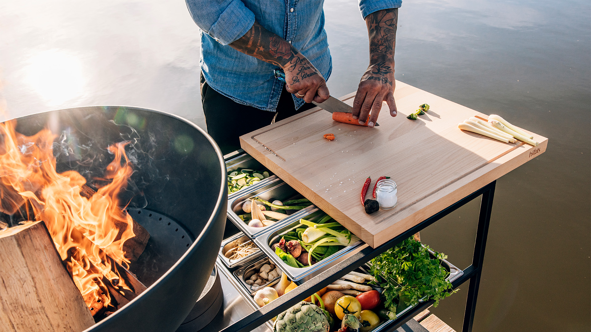 The 00614 Fire Kitchen Worktop integrates seamlessly into the setting, where a man cooking food on a grill on a lake.
