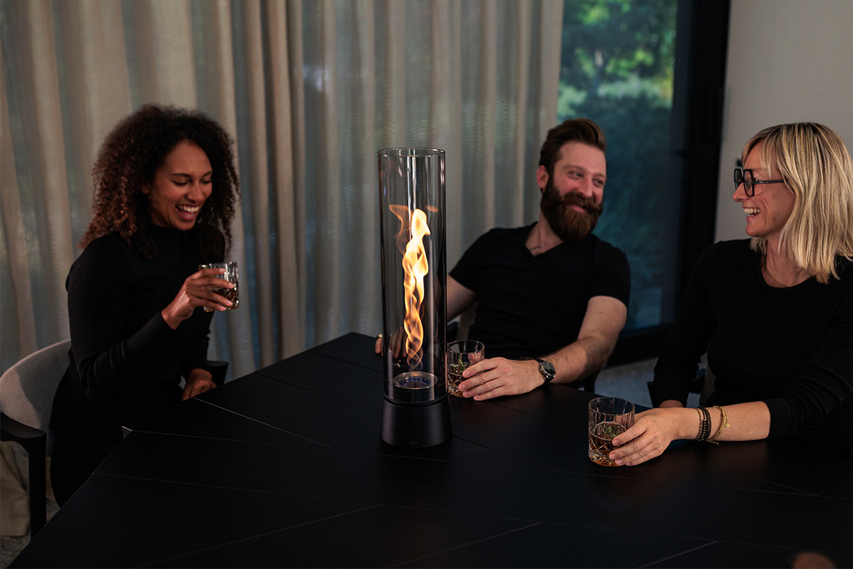 SPIN air 1200 Tabletop Fireplace all black Drei schwarz gekleidete Personen sitzen um SPIN air mit Flamme und amüsieren sich.