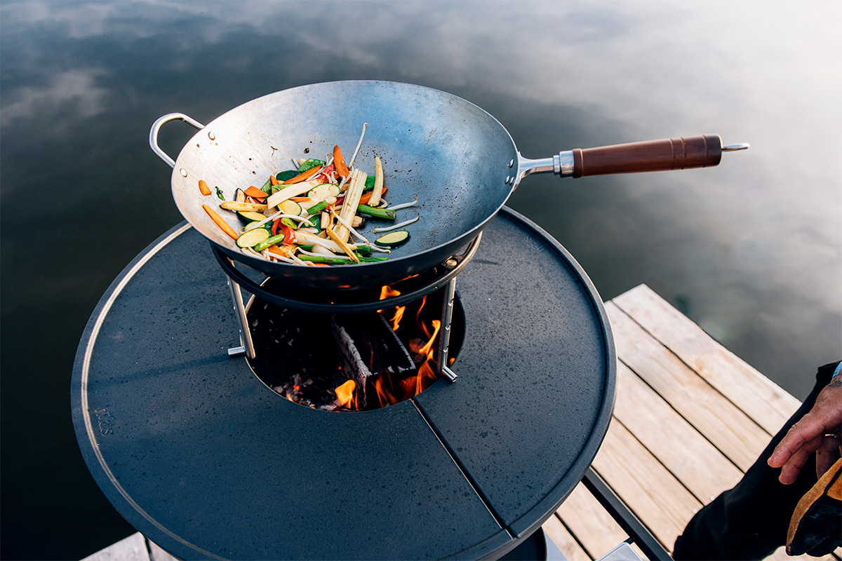 BOWL 70 Sear Grate Depicted here is a person cooking food on a grill on a dock, with the BOWL 70 Sear Grate.