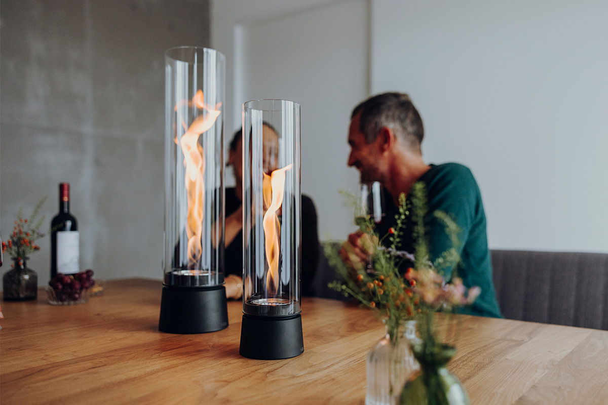 SPIN air 900 Tabletop Fireplace matte black SPIN air 1200 und 900 mattschwarz auf einem Küchentisch mit zwei Personen welche mit einem Rotwein anstoßen und sich unterhalten