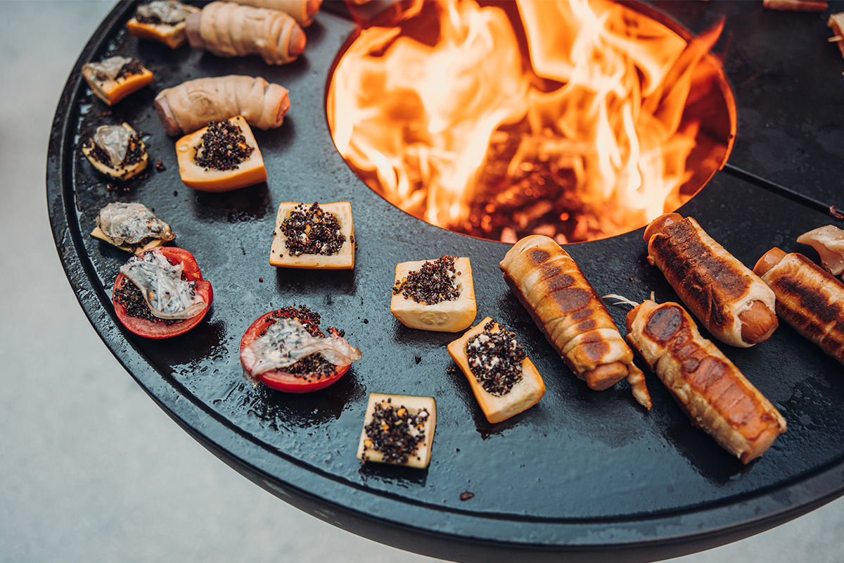 BOWL 57 Plancha BBQ Set low An image showing a fire pit with a bunch of sausages and meat on it, with the BOWL 57 Plancha BBQ Set low.